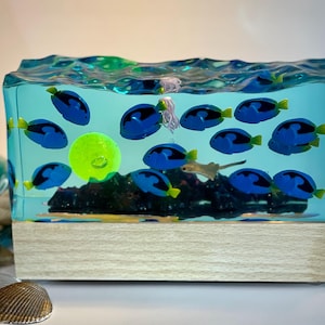 Puede incluir: Lámpara rectangular de resina con una escena submarina. Muestra peces azules con aletas amarillas, una esfera verde y una medusa, todo dentro de una resina transparente de color turquesa. La base es de madera clara.