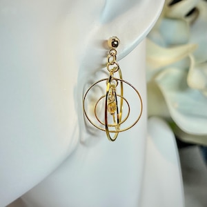 May include: Gold-colored earrings with a unique design. The earrings feature a small gold ball at the top, connected by a chain to a series of concentric circles and a small, dangling charm. The design is geometric and modern, suitable for everyday wear or special occasions.