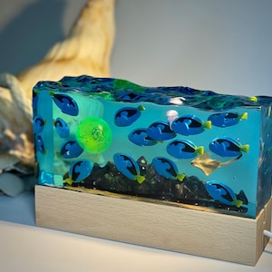 Puede incluir: Lámpara rectangular de resina con peces azules, un orbe verde y una raya, sobre un fondo azul. La lámpara se asienta sobre una base de madera clara, con un cable de alimentación.