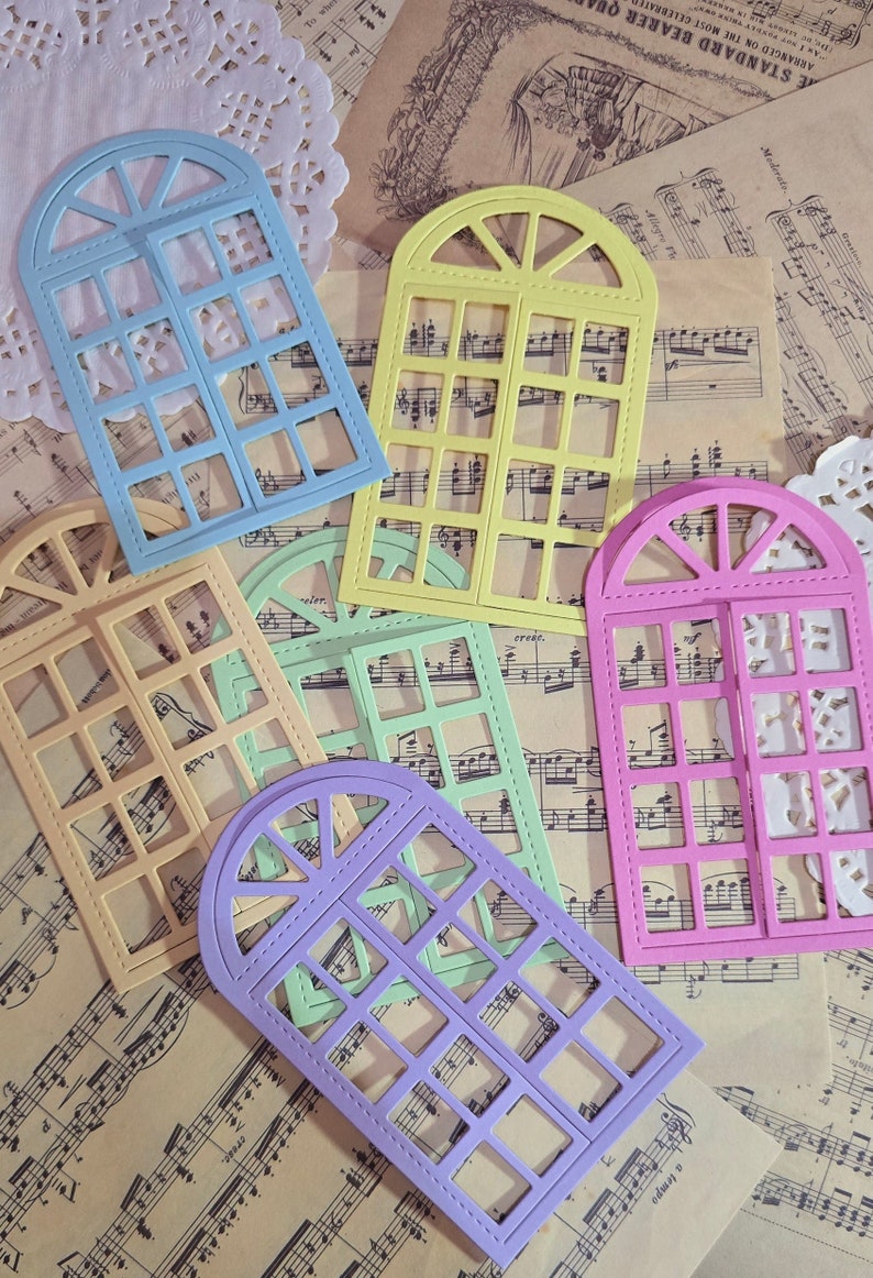 Puede incluir: Cinco troqueles decorativos de marco de ventana en diferentes colores. Los troqueles est&aacute;n dispuestos en una hoja de papel de m&uacute;sica. Los colores son azul claro, amarillo, verde claro, morado y rosa.