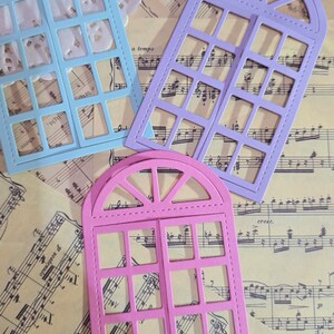 Puede incluir: Tres troqueles decorativos de marco de ventana en azul, morado y rosa. Los troqueles est&aacute;n sobre una hoja de m&uacute;sica.
