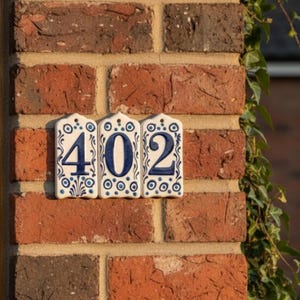 Pode incluir: Três azulejos brancos de cerâmica com o número da casa 402 em azul. Os azulejos são decorados com padrões florais azuis e estão montados numa parede de tijolo vermelho. Os números estão numa fonte arrojada e fácil de ler.