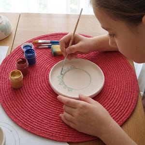 Op de afbeelding: Een persoon beschildert een witte keramische plaat met een bloemmotief met een dunne penseel. Verschillende verfkleuren, waaronder blauw, rood en goud, zijn zichtbaar in kleine potjes. De plaat ligt op een rode geweven placemat.
