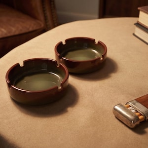 以下が含まれることがあります： 丸いデザインの茶色と緑色のセラミック灰皿が2つ。 灰皿は濃い茶色の外側と緑色の内側をしています。 銀色の金属製のキャップが付いた木製のタバコケースもテーブルにあります。