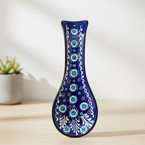 May include: A blue ceramic spoon rest with a black border. The spoon rest is decorated with white and blue eye-shaped designs and floral patterns. The spoon rest is on a wooden surface.