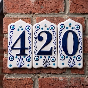 Peut inclure: Trois carreaux de numéro de maison en céramique blanche affichant le numéro "420". Chaque carreau a une bordure décorative bleue et blanche avec des motifs en forme d'œil et floraux. Les chiffres sont en gras, bleu foncé.