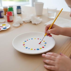 May include: A white ceramic plate being painted with a yellow-handled paintbrush. The plate has a circle of colourful dots in red, yellow, and blue. Paint supplies, including small bottles of paint and a palette, are in the background.