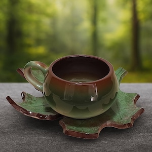 Może przedstawiać: Ceramiczny zestaw filiżanki i spodka. Filiżanka jest okrągła z uchwytem i gradientem od brązu do zieleni. Spodek ma kształt liścia, z podobną zielono-brązową kolorystyką. Zestaw ma przypominać liść.
