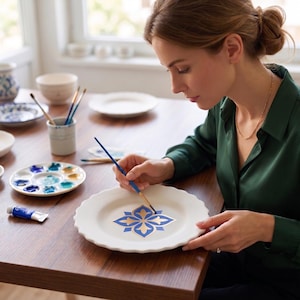 May include: A white ceramic plate with a scalloped edge featuring a hand-painted blue and gold floral motif. The plate is being decorated with a fine-tipped paintbrush, with a paint palette and a tube of blue paint nearby on the table.
