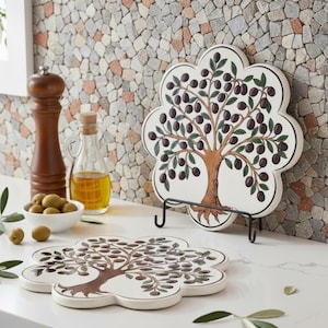 Può includere: Due sottopentola in ceramica bianca con un design floreale raffigurante un albero di ulivo con olive nere e foglie verdi. Un sottopentola è su un supporto in metallo nero. Un macinapepe in legno, olio d'oliva e olive sono anche nell'immagine.