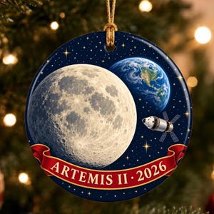 Può includere: Un ornamento rotondo in ceramica con uno sfondo spaziale blu scuro stellato, una grande luna dettagliata, una vista della Terra e una navicella spaziale. Un nastro rosso nella parte inferiore mostra il testo bianco ARTEMIS II 2026 con un cordino dorato.