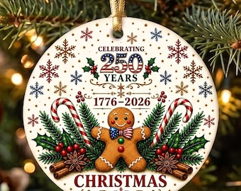 May include: A round white ceramic Christmas ornament featuring a gingerbread person wearing a stars and stripes bowtie, flanked by candy canes, pine branches, and cinnamon sticks. Text reads Celebrating 250 Years 1776-2026 Christmas with snowflakes.