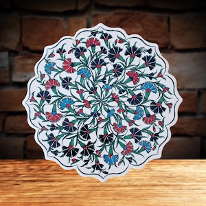 May include: A white, scalloped-edge decorative plate with a floral design. The intricate pattern features red, blue, and black flowers with green stems and leaves. The plate is set on a wooden surface against a brick background.