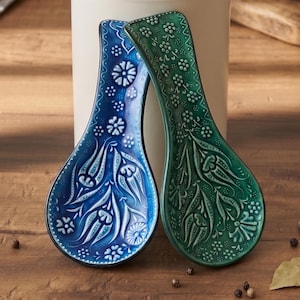 Op de afbeelding: Twee keramische lepelsteunen. Eén is diepblauw met witte bloempatronen, de andere is groen met witte bloempatronen. Beide hebben een druppelvorm en zijn op een houten oppervlak geplaatst.