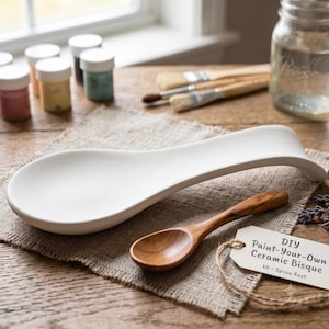 Könnte beinhalten: Eine weiße Löffelablage aus Keramik mit glatter, matter Oberfläche, begleitet von einem kleinen Holzlöffel und einem weißen Papieranhänger mit dem Text DIY Paint-Your-Own Ceramic Bisque k5 - Spoon Rest, auf einem strukturierten braunen Stoff.