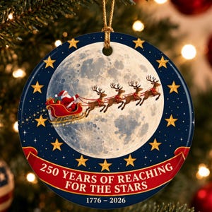Op de afbeelding: Een rond keramisch ornament met een volle maan op de achtergrond en de Kerstman in een rode slee getrokken door vier rendieren. De donkerblauwe rand toont gouden sterren en een rode banner met de tekst 250 YEARS OF REACHING FOR THE STARS 1776 – 2026.