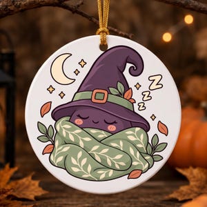 May include: A round white ceramic ornament hanging from a gold cord. The illustration features a purple witch hat with a green band, a green scarf with a white leaf pattern, a crescent moon, stars, and Zs indicating sleep on a white background.