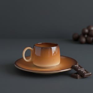 May include: A brown ceramic coffee cup and saucer set. The cup has a rounded shape with a handle, and sits on a matching saucer. Two pieces of dark chocolate are placed next to the set. The background is a dark grey.