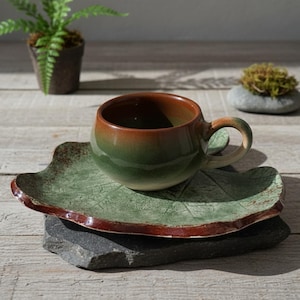 以下が含まれることがあります： 手作りのセラミックティーカップとソーサーのセット。 カップは茶色から緑へのグラデーションで、お揃いのハンドルが付いています。 葉の形をしたソーサーは緑色で、茶色の縁があります。 セットは濃い灰色の石の上に展示されています。