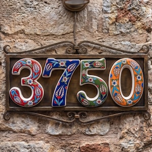 May include: Address plaque displaying the numbers 3750. The ceramic numbers feature floral designs in red, blue, green, and orange. The plaque is mounted on a weathered stone wall.