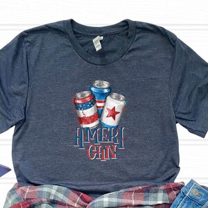 Puede incluir: Una camiseta azul marino con un gráfico de tres latas rojas, blancas y azules con temática de la bandera estadounidense. El texto "Ameri-Can" está impreso debajo de las latas.