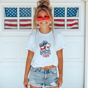Puede incluir: Una camiseta blanca con un gráfico de tres latas rojas, blancas y azules con el texto "Ameri-Can" impreso en la parte delantera.