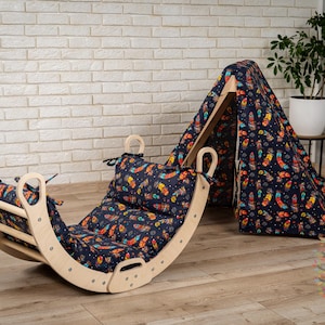 Puede incluir: Un columpio de madera con un cojín estampado de colores. El columpio está diseñado para niños y forma parte de un conjunto de juegos. El cojín tiene un patrón de plumas de varios colores sobre un fondo azul oscuro.