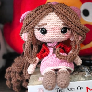 以下が含まれることがあります： 手作りの編み人形。茶色の髪と大きな黒い目。人形はピンクのドレスと赤いショールを着ています。人形は本の上に座っています。