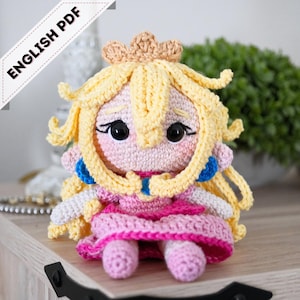 May include: A handmade crochet doll of a princess, featuring a yellow crown and hair, a pink dress, and blue accents. The doll has black eyes and a sad expression. The text "ENGLISH PDF" is in the upper left corner.