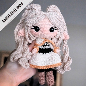 May include: A handmade crochet doll with long, light blonde hair and large black eyes. The doll is dressed in a white dress with a black and white bodice and tan details. The text "ENGLISH PDF" is visible.