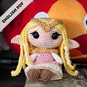 May include: A handmade crochet doll with blonde hair, wearing a pink dress and a tan scarf. The doll has large black eyes and is posed in front of a red background. The text "ENGLISH PDF" is in the upper left corner.