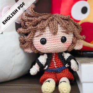 Pode incluir: Boneca de crochê feita à mão com cabelo castanho, olhos pretos e bochechas rosadas. A boneca usa uma roupa vermelha com jaqueta preta e branca e sapatos amarelos. O texto "ENGLISH PDF" é visível. Item feito à mão.