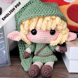 Op de afbeelding: Handgemaakte gehaakte pop van een videogame-personage. De pop heeft een groene tuniek en hoed, blond haar en grote zwarte ogen. De tekst "ENGLISH PDF" staat linksboven. De pop is ongeveer 15 cm groot.