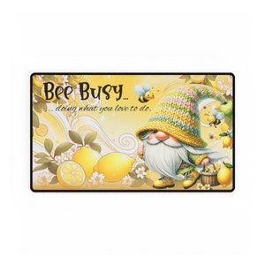 May include: A yellow and green gnome with a white beard and a yellow knitted hat with pink and green stripes. The gnome is holding a basket of lemons and there are bees flying around him. The text "Bee Busy... doing what you love to do." is written in black on a yellow background.