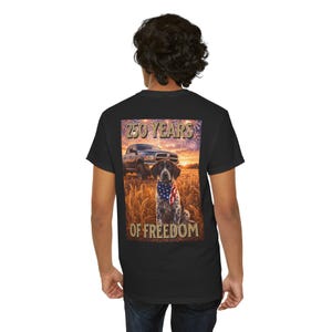 Puede incluir: Camiseta negra con un diseño de un perro con un pañuelo con la bandera estadounidense, una camioneta y el texto "250 YEARS OF FREEDOM". El diseño está ambientado en un campo de trigo y fuegos artificiales.