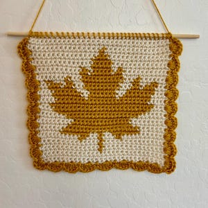 Peut inclure: Une tenture murale carrée en crochet présentant un motif de feuille d'érable jaune moutarde sur fond crème. La pièce est bordée d'une bordure festonnée jaune moutarde et est suspendue à une tige en bois horizontale avec une ficelle assortie.