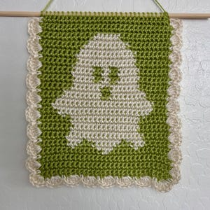Könnte beinhalten: Ein rechteckiger Wandbehang aus Häkelarbeit mit einem weißen Geistermotiv auf grünem Hintergrund. Das Stück ist mit einer weißen, bogenförmigen Bordüre eingefasst und hängt an einem horizontalen Holzstab, der mit einer grünen Schnur befestigt ist.