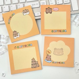 Peut inclure: Quatre blocs-notes orange sur le thème du Capybara avec des illustrations de capybaras et le mot "CAPYBARA". Une note a une forme ronde et une autre une forme de nuage. Les notes sont sur un clavier blanc.