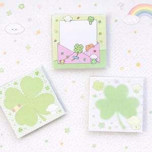 May include: Three square sticky note pads with a clover and cartoon design. One pad features a four-leaf clover, another has a clover design with an envelope, and the third has a clover outline. The pads are light green and white.