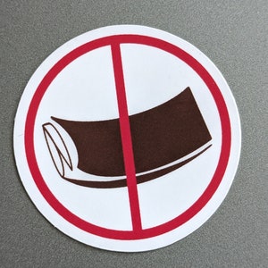 May include: A round white sticker with a red border and a red diagonal line through the center. Inside the circle is a brown object, possibly a rolled-up item, with a white outline. The sticker is on a gray surface.