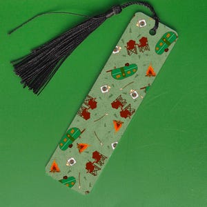 May include: A green bookmark with a black tassel. The bookmark features a repeating pattern of camping elements, including tents, campfires, and camping chairs. The bookmark is approximately 7 inches long.