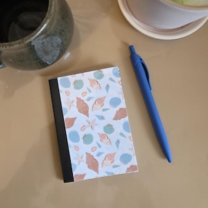 Peut inclure: Un petit carnet avec une reliure noire et une couverture à motifs de coquillages dans des tons pêche, bleu et vert sur fond blanc. Un stylo bleu est posé à côté du carnet sur une surface beige. Une tasse en céramique est dans le coin supérieur gauche.