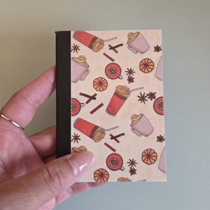 Könnte beinhalten: Ein kleines Notizbuch mit schwarzem Rücken und cremefarbenem Einband mit einem sich wiederholenden Muster aus Kaffeegetränken, Zimtstangen und Orangenscheiben. Das Notizbuch wird von einer Hand gehalten.