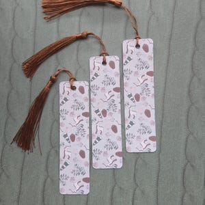 May include: Three rectangular bookmarks with a light purple background and a repeating pattern of brown pinecones and other shapes. Each bookmark has a brown tassel attached to the top with a string.