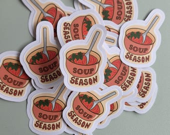 May include: A pile of stickers depicting a bowl of soup with a spoon. The stickers have a light orange border and a light orange bowl with red soup and green garnish. The text "SOUP SEASON" is printed on the stickers.