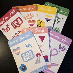 Pode incluir: Uma coleção de autocolantes coloridos com o tema do Orgulho, com vários desenhos e texto. Os autocolantes apresentam frases como "Kiss Me I'm Gay" e "Let Me Be Perfectly Queer". Os autocolantes estão dispostos numa superfície escura.