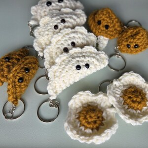 May include: A collection of handmade crochet keychains in various shapes. The keychains include white dumplings, brown cookies, and a white flower with a yellow center. Each keychain has a silver ring and black bead eyes.