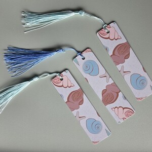 Op de afbeelding: Vier rechthoekige bladwijzers met schelp- en zeesterpatronen in roze, blauw en bruin. Elke bladwijzer heeft een kwast in blauwe en witte tinten. De bladwijzers liggen op een lichtgrijze ondergrond.