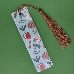 May include: A light blue bookmark with a brown tassel. The bookmark features a repeating pattern of cocoa mugs, chocolate bars, and sprigs. The tassel is made of brown string.