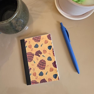 Op de afbeelding: Een klein notitieboekje met een zwarte rug en een patroon op de kaft met geruite shirts, hoeden en andere vormen in tinten bruin, blauw en paars op een gele achtergrond. Een blauwe pen ligt ernaast op een beige oppervlak.